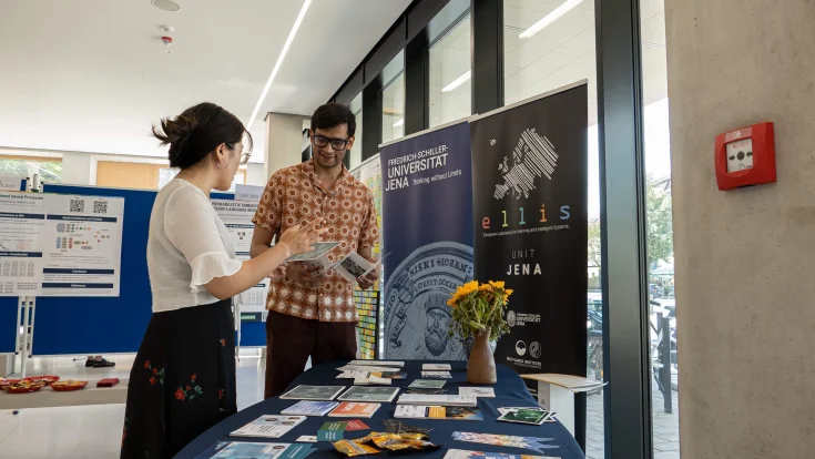 Two people talking at a table with flyers at ELLIS Unit Jena