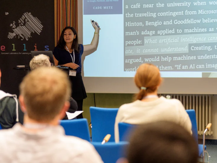 a woman is pointing at a screen and presenting in front of several people sitting in chairs