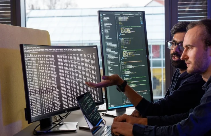 Two men looking at 2 screens with computer code on them
