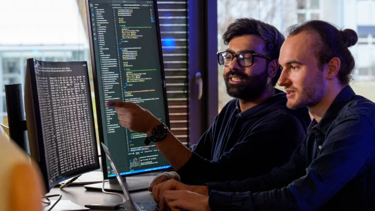 Two men sitting in front of 2 large screens with computer code on them