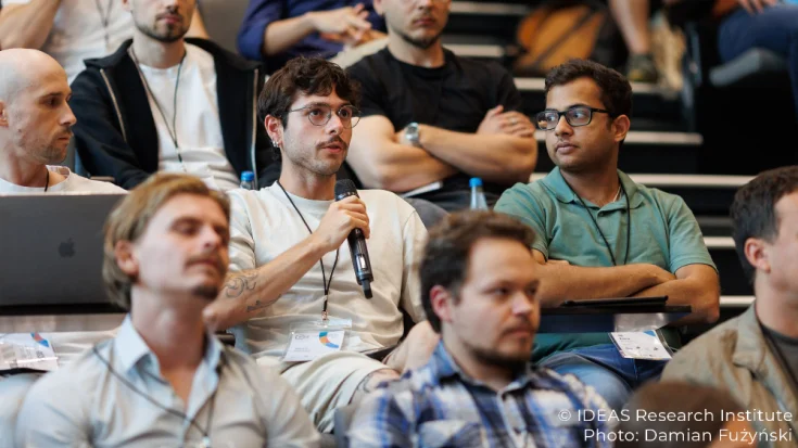 An ELLIS PhD student speaks into a microphone while several other