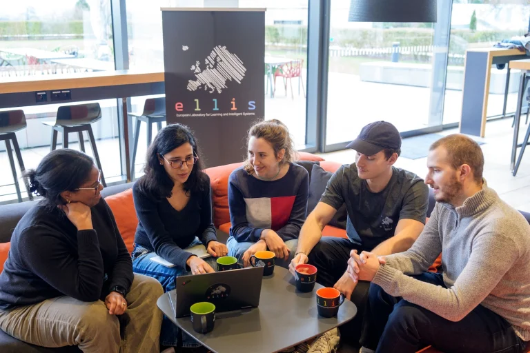 Group of people sitting around a coffee table with ELLIS mugs, looking at one another and at a laptop, with an ELLIS rollup in the background